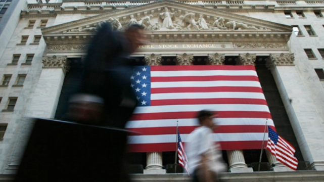 A series by CJS alumni covering the roots and impact of several corporate scandals that became symbolic of the modern gilded age (Ruth Fremson/The New York Times). Street-level view of the New York Stock Exchange building, prominently displaying a large American flag on its facade.