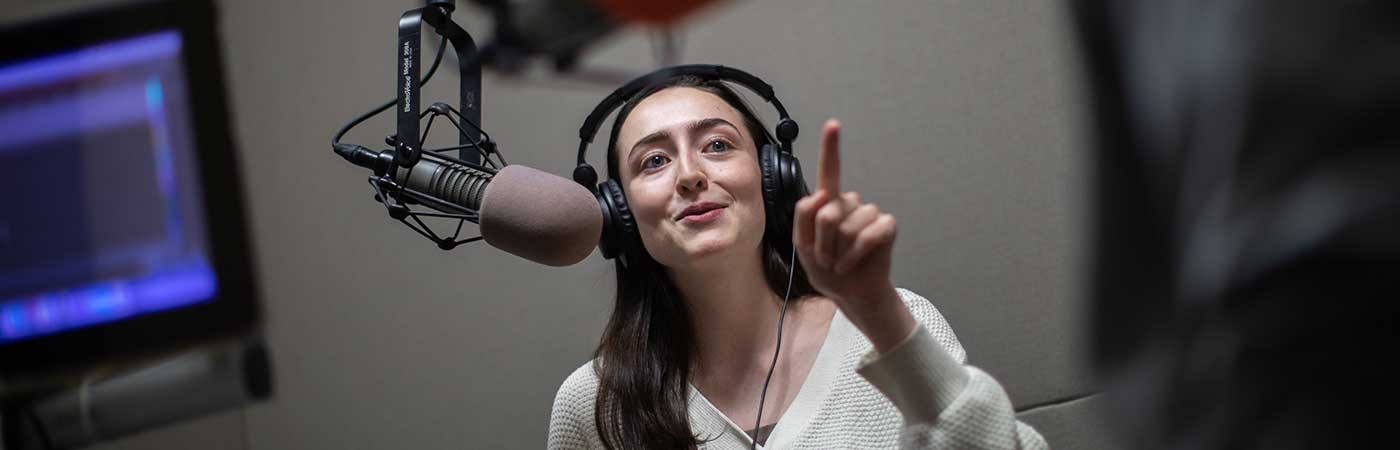 female student in radio studio wearing headphones and gesturing towards another participant in a discussion