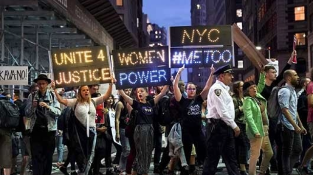 Protesters march through Midtown Manhattan as they rally against Supreme Court nominee Judge Brett Kavanaugh, Oct. 1, 2018 in New York. Photo by Drew Angerer/Getty Images. Group protests holding lit up signs that say, "Unite 4 Justice," "Women Power," and "NYC #MeToo."