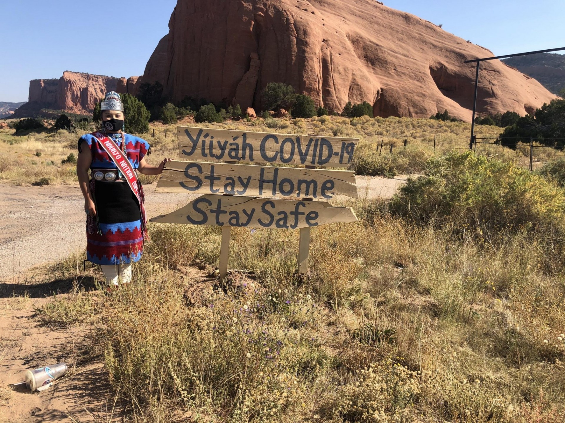 Navajo woman in facial mask, crown and dress stands next to sign readiing "Stay Safe, Stay Home" with mesa formation behind her