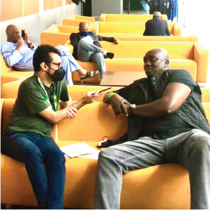 Two men on a yellow couch. One is holding a phone as he interviews the other.