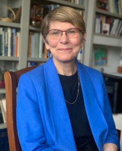 Women wearing glasses and a blue blazer sits in a wooden chair in front of a bookshelf. 