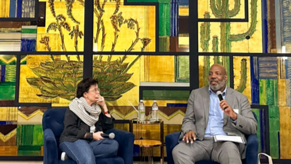 A two-person panel with a woman on the left and a man on the right, sitting in front of a stained glass wall. The vibrant colors of the stained glass create a striking backdrop behind them.