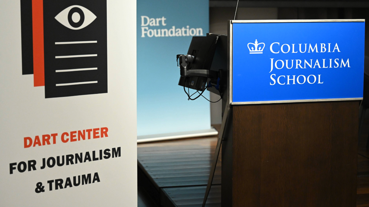 Two banners and one podium on a stage. The far left banner has a white background and reads "Dart Center for Journalism and Trauma" with a logo featuring an eye on three pieces of stacked paper, which are black and red. The middle banner reads "Dart Foundation" in white font on a light blue background. The podium on the far right is blue with white font and features the Columbia crown logo, reading "Columbia Journalism School."