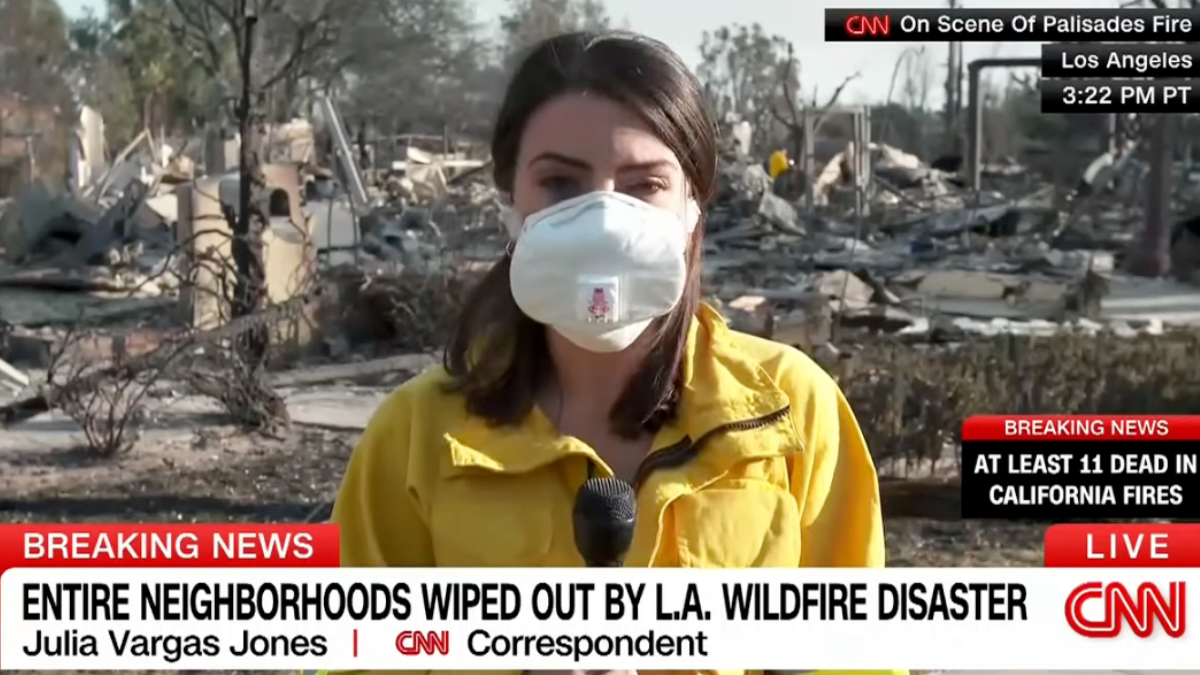 A woman wearing a yellow jacket stands in front of debris during a CNN broadcast.