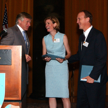 Mary Jordan, ‘84 M.S., accepts the 2003 Pulitzer Prize in International Reporting. Photo: The Pulitzer Prizes. From left to right, a man, a woman, and another man are on stage. The woman is accepting an award while the two men stand beside her.