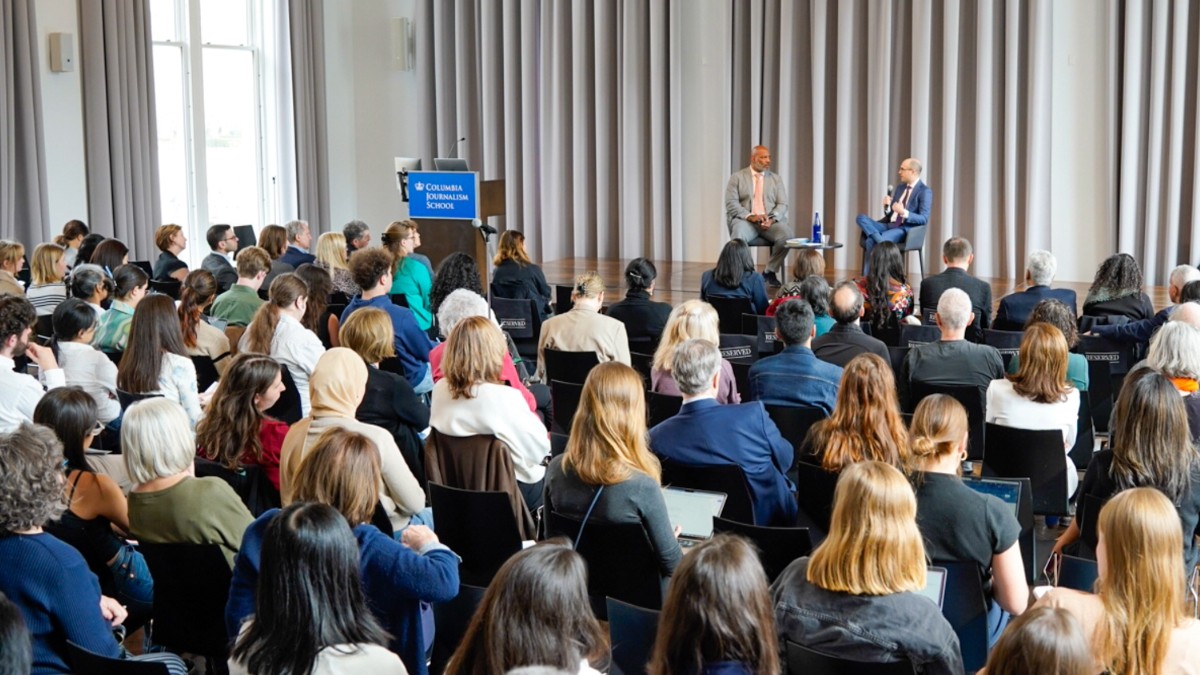 A crowd faces a stage featuring a two-person panel. 