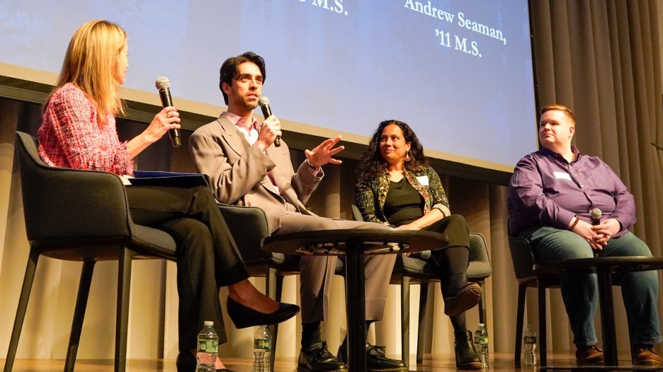 A four-person panel on a stage. Each panelist is holding a microphone, one is actively speaking. 