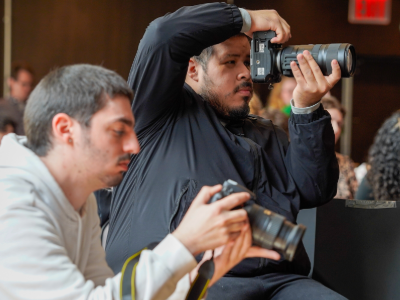 Two photographers at an event: one is actively taking a photo with their camera, while the other reviews an image on their camera’s screen.