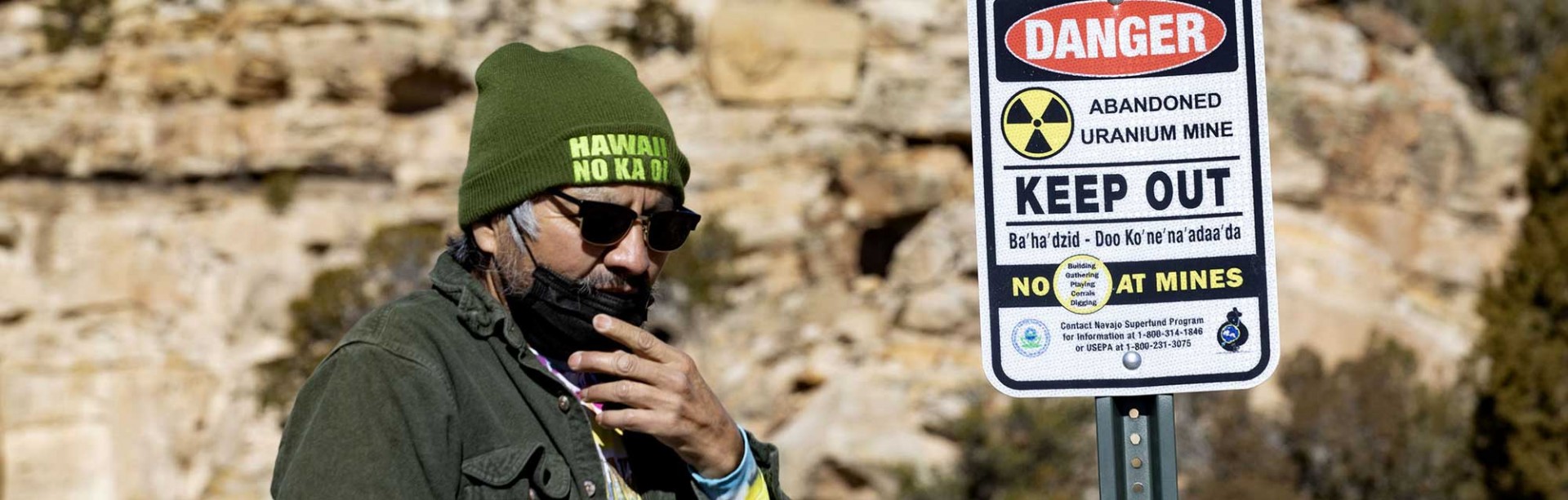Photo of a man in knitted cap and sunglasses outdoors by a sign warning of danger from an abandoned uranium mine