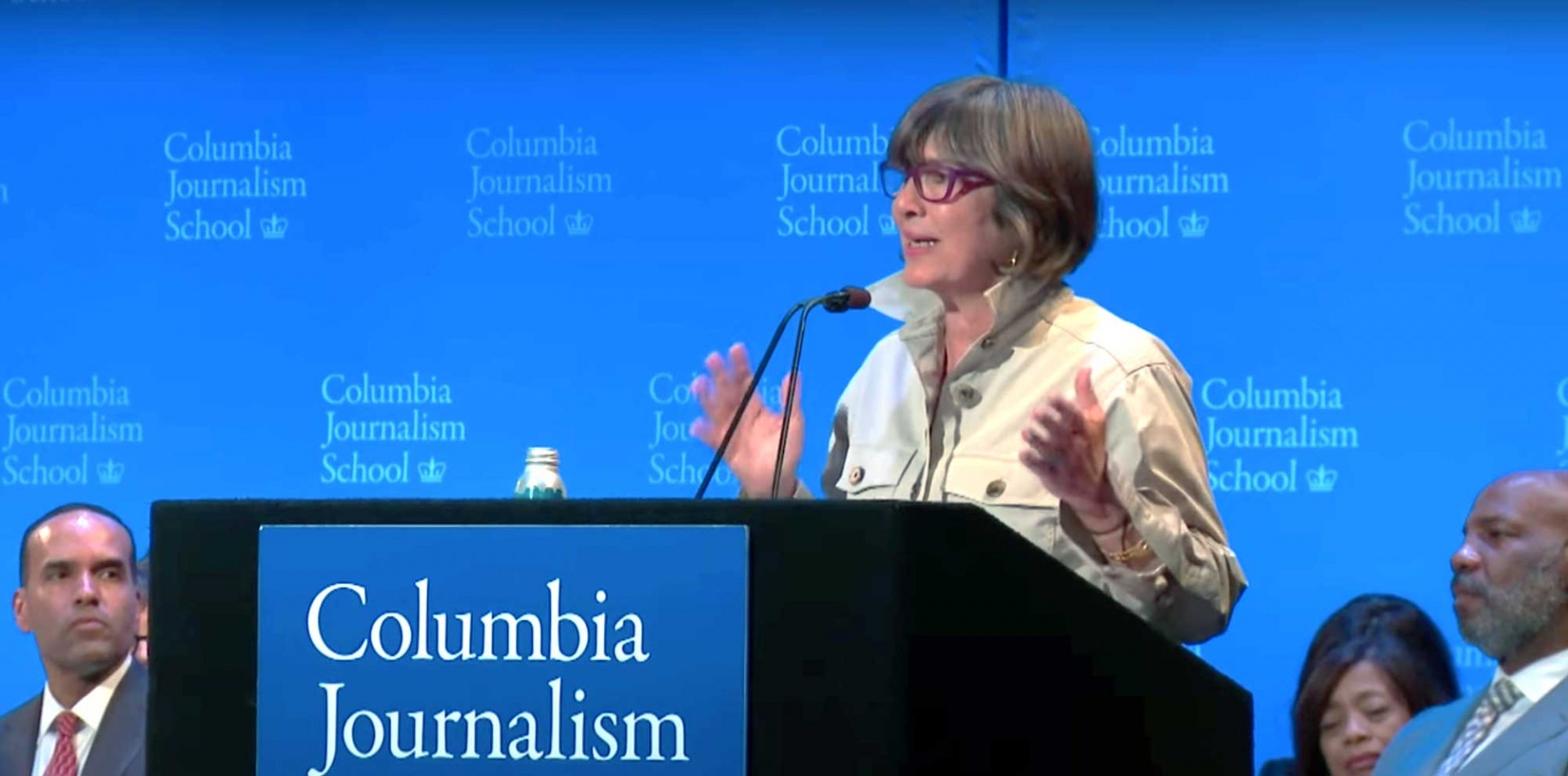 Christiane Amanpour speaks while standing at podium, with CJS Dean of Student Life John Haskins and CJS Dean Jelani Cobb seated on the stage behind her.