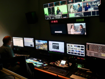 Multiple monitors in a broadcast studio editing room displaying video feeds, editing software, and control panels.