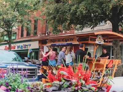 The Sip-and-Cycle, making its way through downtown Sioux Falls. (Rita Omokha/Elle) Flowers in the foreground; people on a "Sip n Cycle" behind.