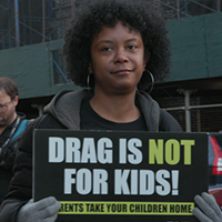 A woman holding a sign that reads, "Drag is not for kids! Parents take your children home," standing in a public space.