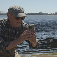 A man wearing a hat and sunglasses takes a photo with a smartphone while standing by the waterfront.
