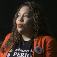 A woman is being interviewed. She is wearing an "alliance for periods" shirt and period-poverty advocacy pins overtop her red blazer.