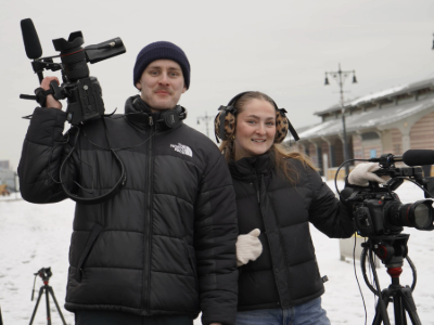 Two students standing together, each holding professional video equipment, including a camera and a boom microphone.