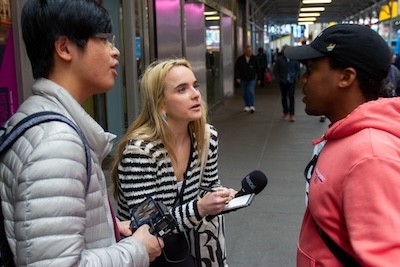 Students in Ty Lawson's City Newsroom class, Vincent Jiang and Ashley Miller, interviewing a local in Times Square. A man with a camera and a woman with a microphone and notepad interview a male pedestrian
