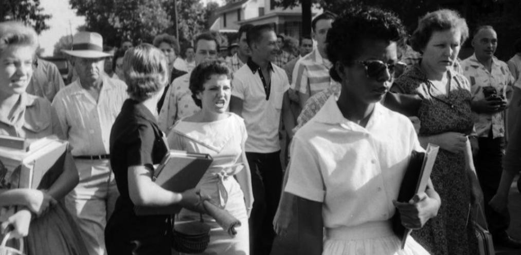 photo of black woman walking ahead of a crowd of white people, with one white woman jeering at the black woman 