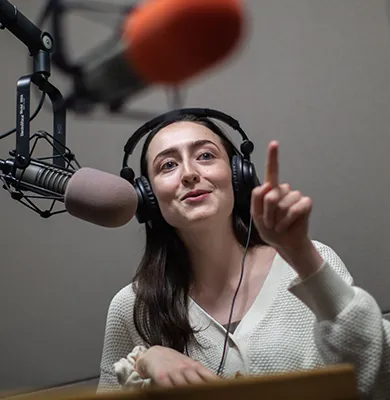 Woman addressing someone during an audio recording session in a recording booth, two microphones.
