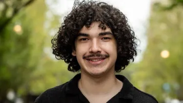 A young person with curly hair and a mustache, wearing a black shirt, smiling outdoors.