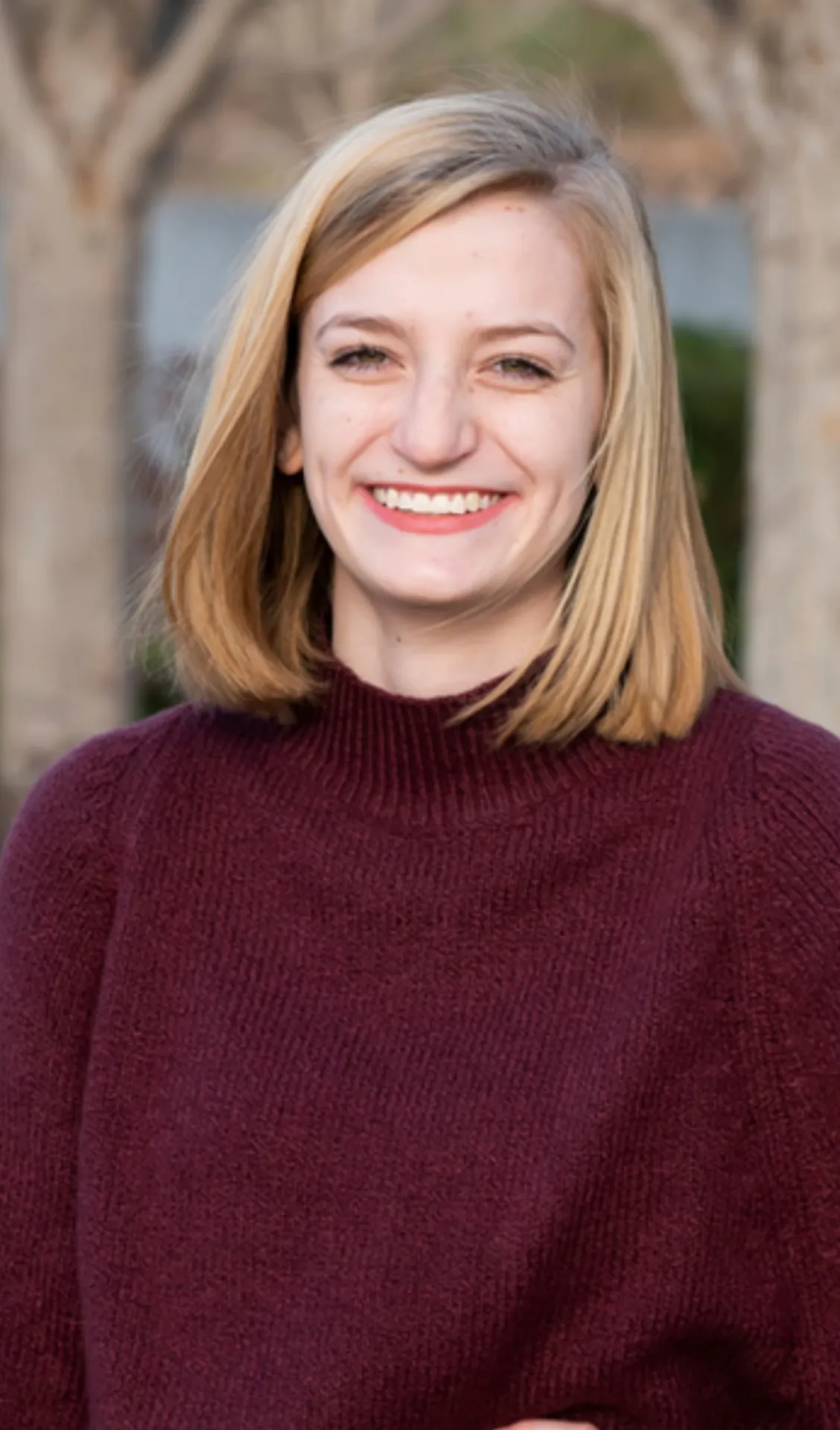 Person with blonde hair smiling, wearing a maroon sweater, outdoors.