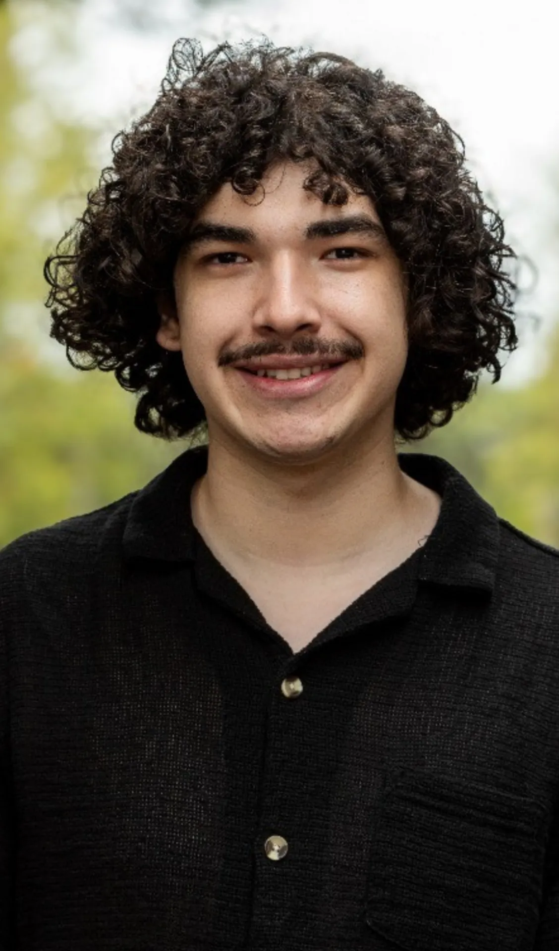 A young person with curly hair and a mustache, wearing a black shirt, smiling outdoors.