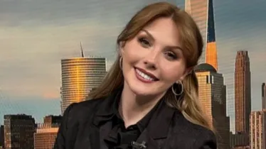 Person seated at a news desk with a city skyline backdrop.