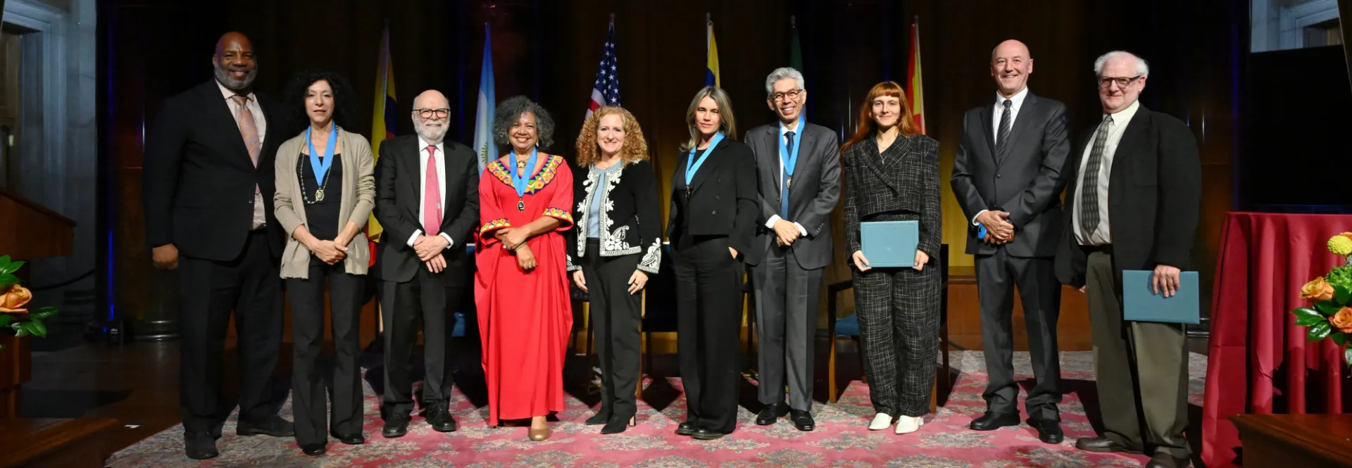 Ten people pose on a stage in front of dark curtains and national flags.