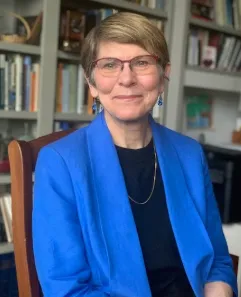 Women wearing glasses and a blue blazer sits in a wooden chair in front of a bookshelf. 