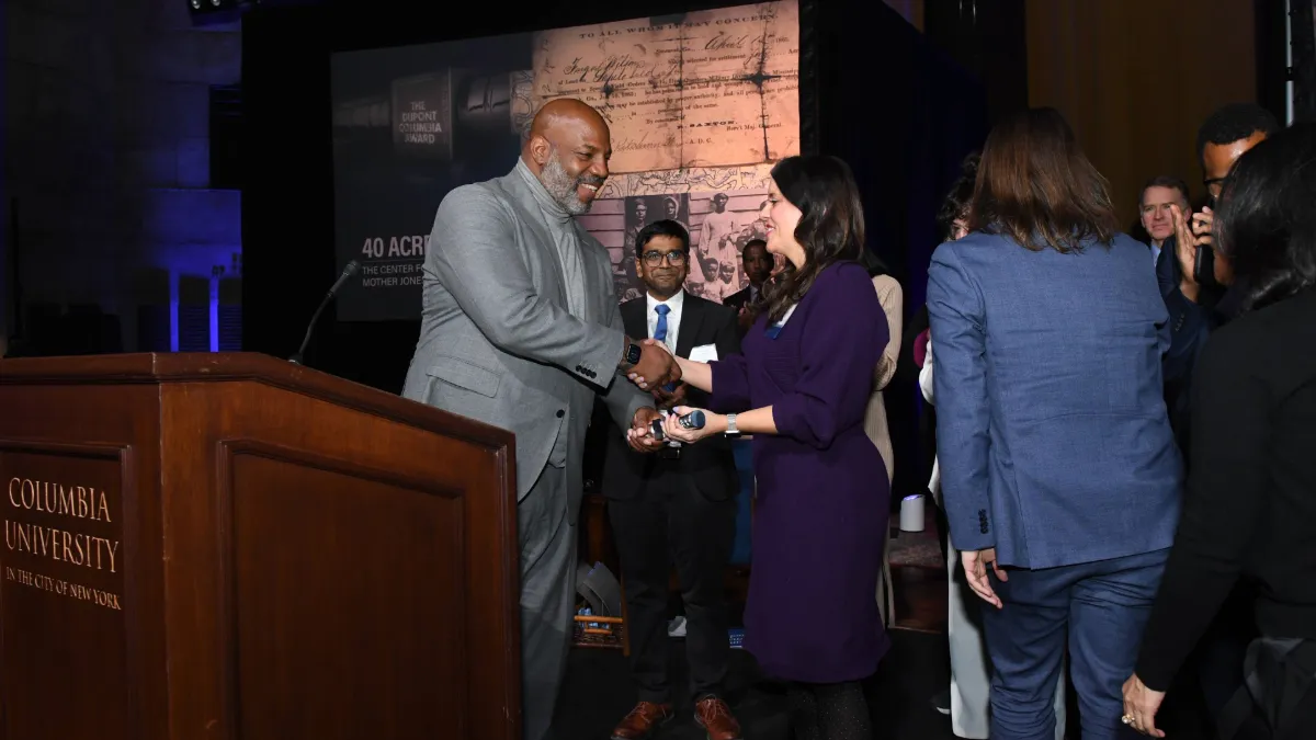 Dean Jelani Cobb stands near a podium and hands the silver baton award to Alexia Fernández Campbell