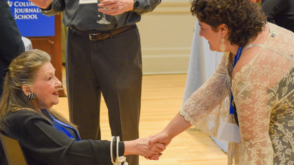 Two women shaking hands, one is sitting (left) the other is standing (right). 