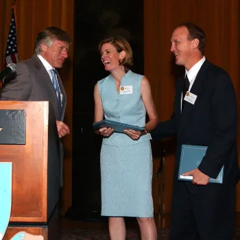 From left to right, a man, a woman, and another man are on stage. The woman is accepting an award while the two men stand beside her.