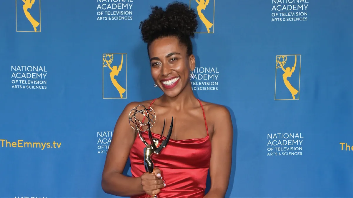Cydney Tucker, ‘16 M.S., clad in a red silk-satin dress, is holding an Emmy Award in front of a blue backdrop. 
