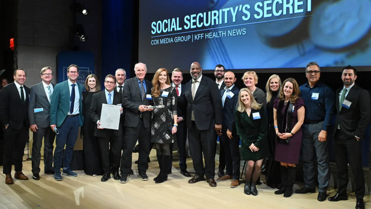A group of sixteen people smiling on a stage holding a certificate and trophy, with a screen displaying "SOCIAL SECURITY'S SECRET" in the background.