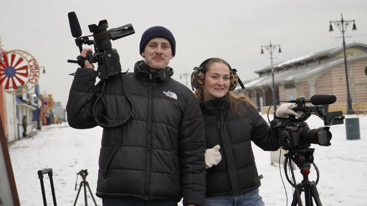 Two students with camera equipment with Coney Island as the backdrop. 