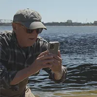 A man wearing a hat and sunglasses takes a photo with a smartphone while standing by the waterfront.