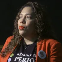 A woman is being interviewed. She is wearing an "alliance for periods" shirt and period-poverty advocacy pins overtop her red blazer.  