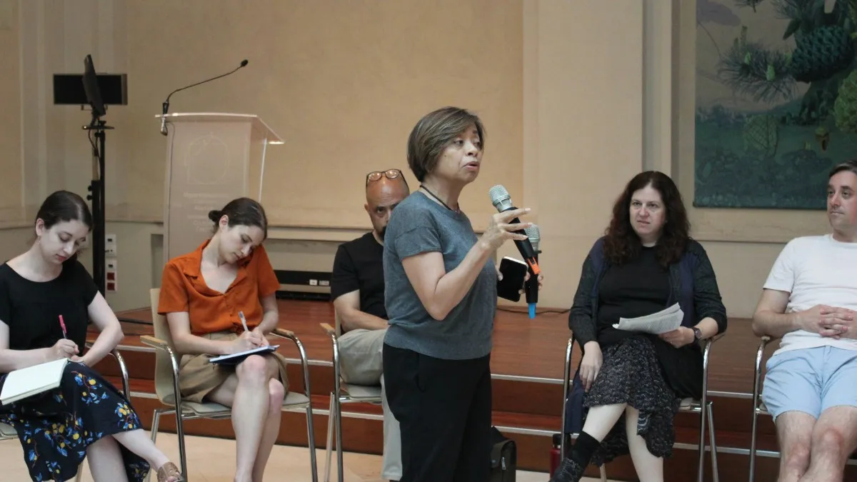 Professor Sheila Coronel speaks into a microphone while others seated nearby are taking notes and listening.