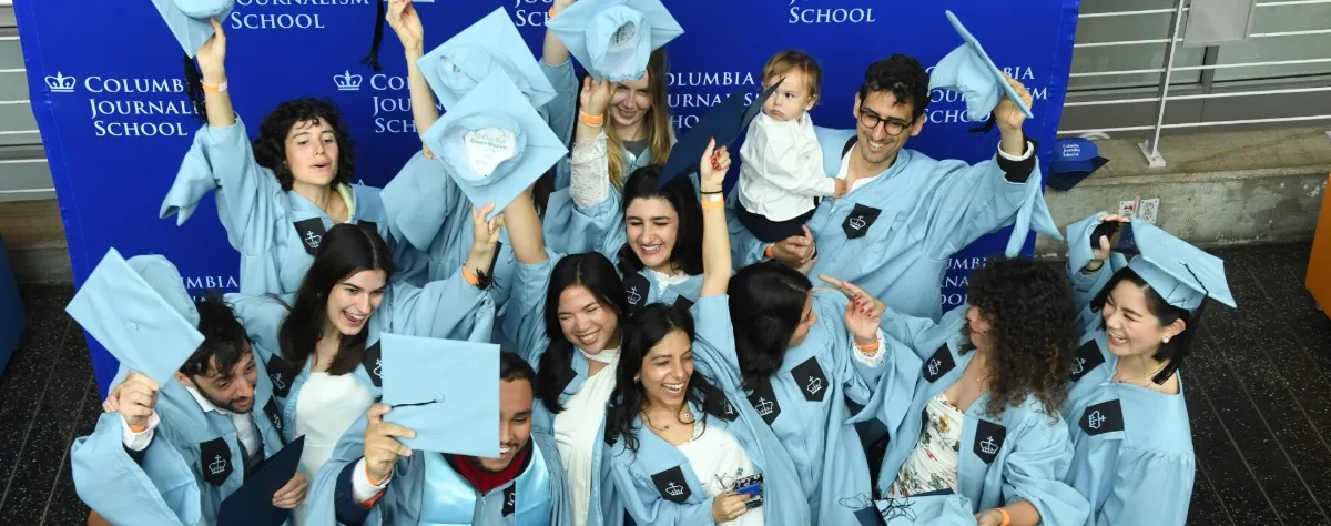 Group of graduates celebrating in light blue gowns in front of a Columbia Journalism School backdrop.
