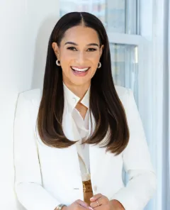 A person smiling in a white blazer stands by a window.