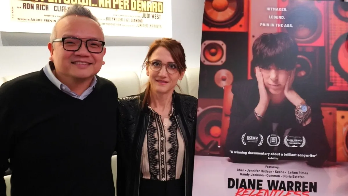 Two people standing indoors next to a poster of a documentary about Diane Warren.