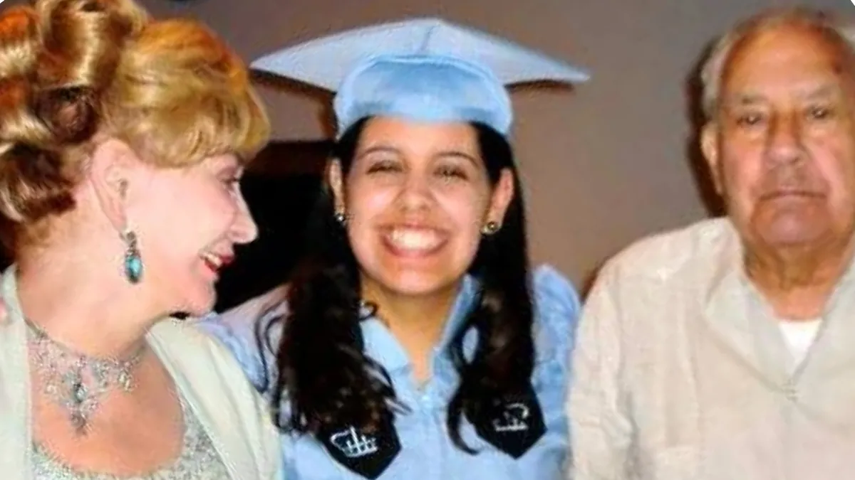 Young woman in a light blue graduation cap and gown smiles between an older woman and an older man.