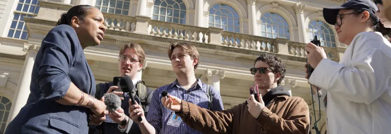 Five people conduct an outdoor interview in front of New York City Hall. 
