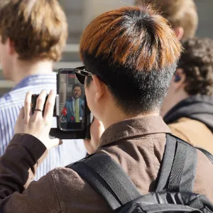 A person records a suited individual on a smartphone while standing among a crowd.