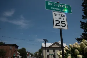 Roadside signs for Bridgewater’s corporate limits and a 25 mph speed limit, with buildings and flowering shrubs under a blue sky.
