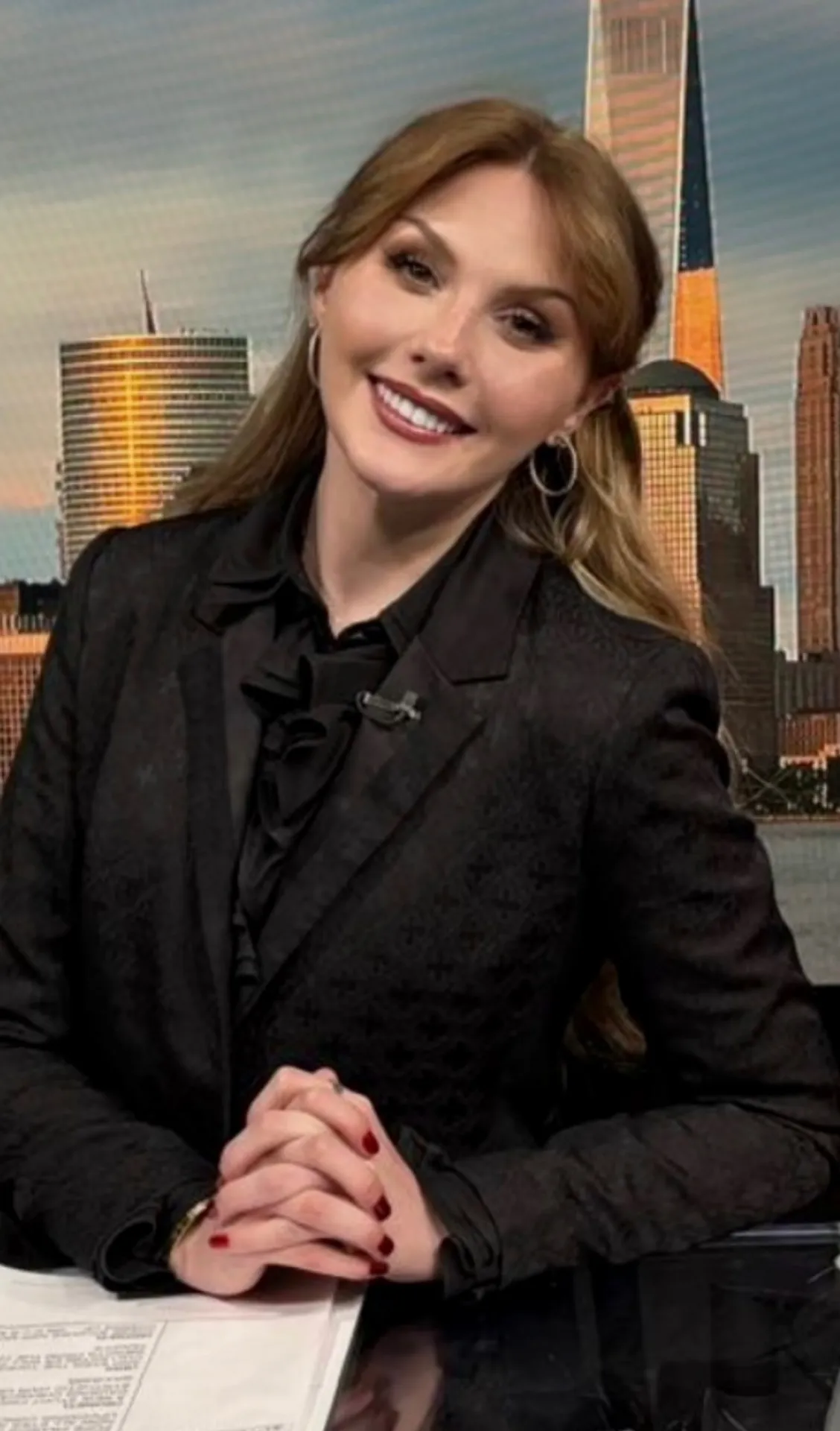 Person seated at a news desk with a city skyline backdrop.